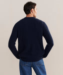 A man stands with his back to the camera, wearing a classic navy Mens Cashmere Fisherman sweater and blue jeans against a plain light background.