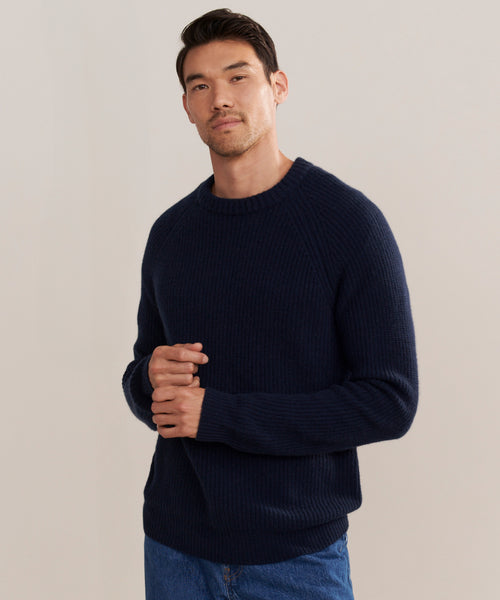 A man with short dark hair wears a navy Mens Cashmere Fisherman sweater and blue jeans against a plain light background, facing forward with a relaxed, neutral expression.