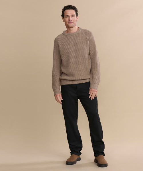 A man with short curly hair models the Men's Cashmere Fisherman Sweater in beige with black pants and brown shoes, standing against an undyed stone background and smiling softly at the camera.