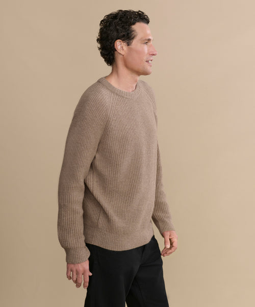 A man with short curly hair, in profile against a plain beige background, wears the Men's Cashmere Fisherman Sweater in undyed stone and black pants.