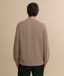 A person with short, curly dark hair is seen from behind wearing the Men's Cashmere Fisherman Sweater in undyed stone and black pants, standing against a plain beige background.