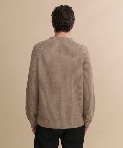 A person with short, curly dark hair is seen from behind wearing the Men's Cashmere Fisherman Sweater in undyed stone and black pants, standing against a plain beige background.
