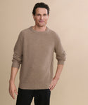 A man with short curly brown hair wears a Men's Cashmere Fisherman Sweater in undyed stone and black pants, standing against a light beige background. He smiles softly, with one hand by his side and the other resting on his hip.