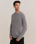 A man wearing the Mens Cashmere Fisherman sweater in heather grey with black pants stands against a plain light background, gazing slightly to the side with a neutral expression.