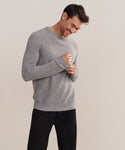 A man wearing a Mens Cashmere Fisherman sweater in light heather grey and black pants stands against a plain background, smiling and looking down with his hands gently clasped.