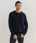 A man with short dark hair, dressed in the Mens Cotton Fisherman sweater and dark jeans, stands against a plain light background, smiling softly with one hand in his pocket.