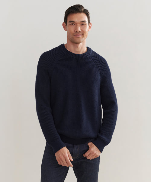 A man with short dark hair, dressed in the Mens Cotton Fisherman sweater and dark jeans, stands against a plain light background, smiling softly with one hand in his pocket.