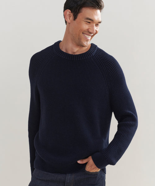 Smiling man wearing a dark blue Mens Cotton Fisherman sweater with ribbed cuffs and collar, standing with one hand in his pocket against a light background. This sweater offers a classic, lightweight knit for everyday comfort.