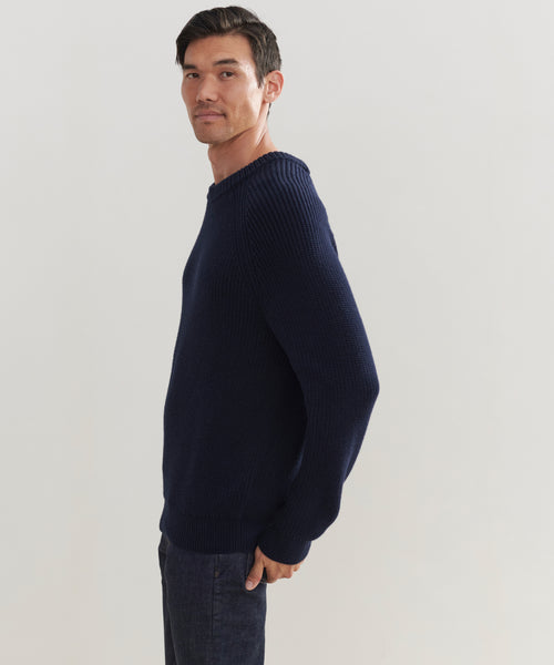 A man stands sideways, looking at the camera, wearing the Mens Cotton Fisherman sweater in navy blue and dark jeans against a plain light background.