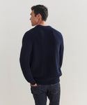 A man with short dark hair, seen from the back, wears a navy blue Mens Cotton Fisherman sweater and dark jeans against a plain light background.