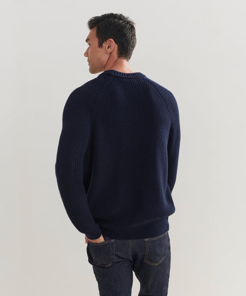 A man with short dark hair, seen from the back, wears a navy blue Mens Cotton Fisherman sweater and dark jeans against a plain light background.