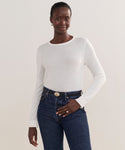 A woman stands against a plain background, wearing the classic white Mercer Crewneck sweater tucked into high-waisted blue jeans with a black belt, posing with one hand in her pocket and a slight smile.