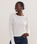 A person with dark skin and short hair is smiling and looking to the side, wearing a white Mercer Crewneck and dark blue jeans against a plain light background.