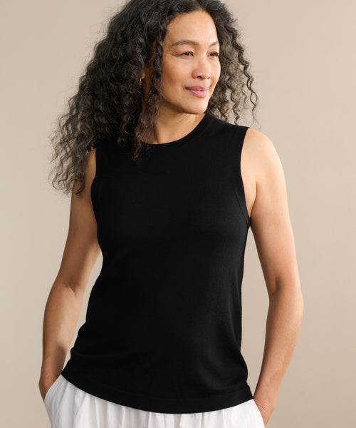 A woman with long, curly hair smiles softly as she stands against a neutral background, hands in her pockets, wearing the Merino Tank and white pants.
