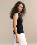 A woman with long, curly hair wears the black Merino Tank and a white skirt, standing sideways and glancing at the camera against a plain light beige background.