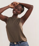 A smiling person with short hair poses with arms raised in a Merino Tank (olive), blue jeans, and black belt against a plain light background.