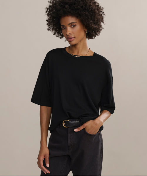 A woman with curly dark hair wears an oversized fit black Merino Tee, dark jeans, a black belt with a gold buckle, a gold necklace, and hoop earrings. She stands against a plain beige background with one hand in her pocket.