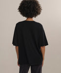Someone with short curly hair, seen from behind, wears an oversized black Merino Tee with dark pants, standing before a plain light background.