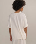 Person with short curly hair wears an oversized Merino Tee in white with matching white pants—an elevated basic, shown from the back against a neutral background.