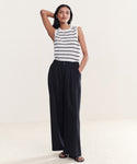In a minimal setting, a woman models the Merino Tank with black stripes, wide-leg black trousers, and black sandals. One hand is in her pocket while the other rests behind her head.