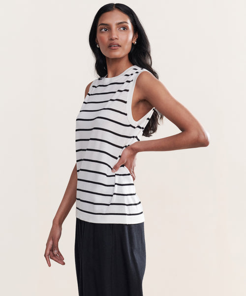 A woman with long dark hair stands sideways against a plain background, wearing the Merino Tank—sleeveless with black horizontal stripes—and black pants, posing with one hand on her hip.