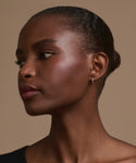 A woman with smooth, dark skin and short, slicked-back hair is shown in profile against a beige background. She wears Merit Baguette Hoops in white gold and a black top, gazing thoughtfully to the side.