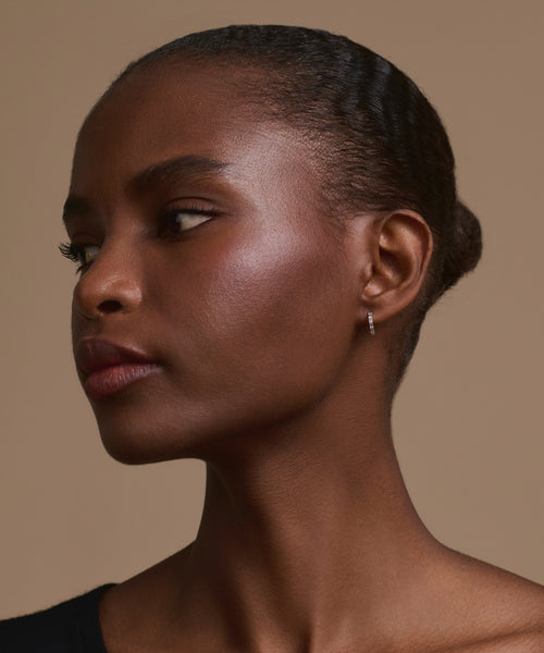 A woman with smooth, dark skin and short, slicked-back hair is shown in profile against a beige background. She wears Merit Baguette Hoops in white gold and a black top, gazing thoughtfully to the side.