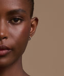Close-up of a woman with smooth brown skin and natural makeup, wearing Merit Baguette Hoops in white gold. Her expression is neutral, set against a beige background.