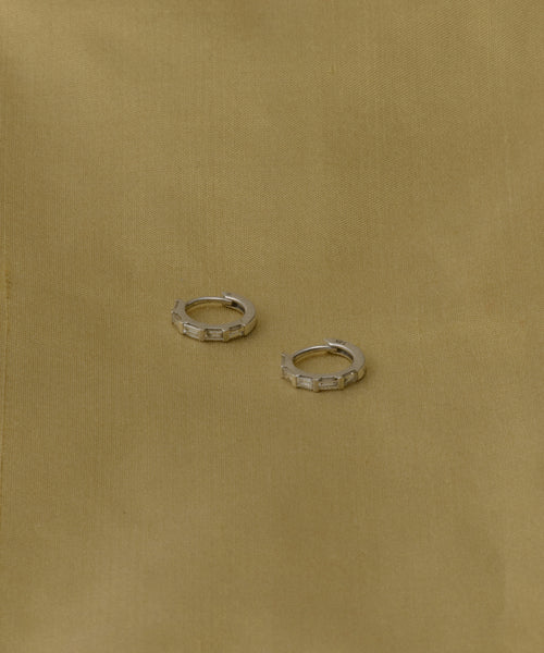 The Merit Baguette Hoops are small white gold earrings with a geometric design, evoking classic heirloom style, displayed on a simple light brown fabric background.