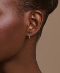 Close-up of a person's ear wearing Merit Baguette Hoops—small, thin white gold earrings with embedded stones for a timeless heirloom feel. The background is neutral beige, showing only part of the face and head.