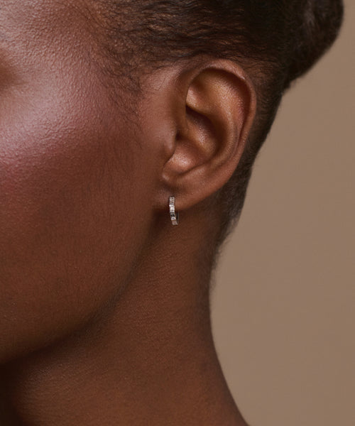 Close-up of a person's ear wearing Merit Baguette Hoops—small, thin white gold earrings with embedded stones for a timeless heirloom feel. The background is neutral beige, showing only part of the face and head.