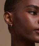 A close-up of a woman's face and ear with smooth brown skin, wearing Merit Baguette Hoops—elegant 14k gold huggies set with tiny stones. The background is a neutral beige color.