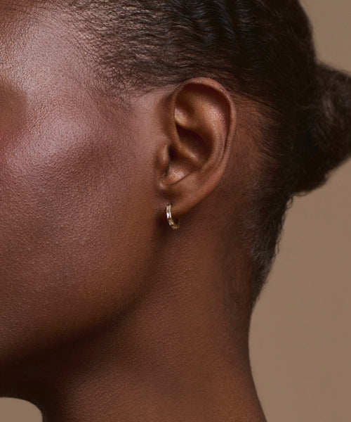 Close-up of a person’s right ear wearing Merit Baguette Hoops in 14k recycled gold. The deep brown skin and pulled-back hair highlight the sleek earrings against a neutral background.