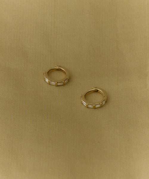 The Merit Baguette Hoops, a pair of small 14k gold huggies with rectangular details, are displayed on textured light brown fabric.