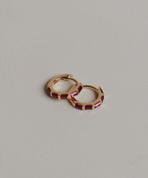 The Merit Baguette Hoops are 14k solid gold earrings with rectangular red gemstones, evoking classic ruby hoop elegance, shown on a light textured background.