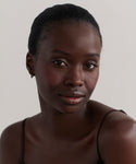 A woman with dark skin and short, pulled-back hair gazes at the camera with a neutral expression, wearing a thin strap top and delicate Merit Baguette Hoops in 14k gold against a light, plain background.