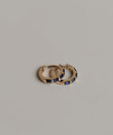 The Merit Baguette Hoops, featuring two small 14k gold hoops with rectangular blue sapphire gemstones, are displayed on a textured light gray surface.