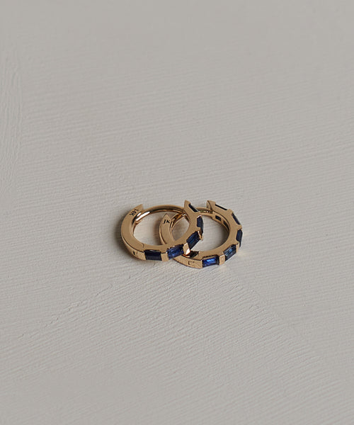 The Merit Baguette Hoops, featuring two small 14k gold hoops with rectangular blue sapphire gemstones, are displayed on a textured light gray surface.