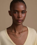 A woman with short hair wears a cream V-neck top and the Merit Chain, a delicate 14k solid gold necklace, while standing against a beige background and looking directly at the camera with a neutral expression.