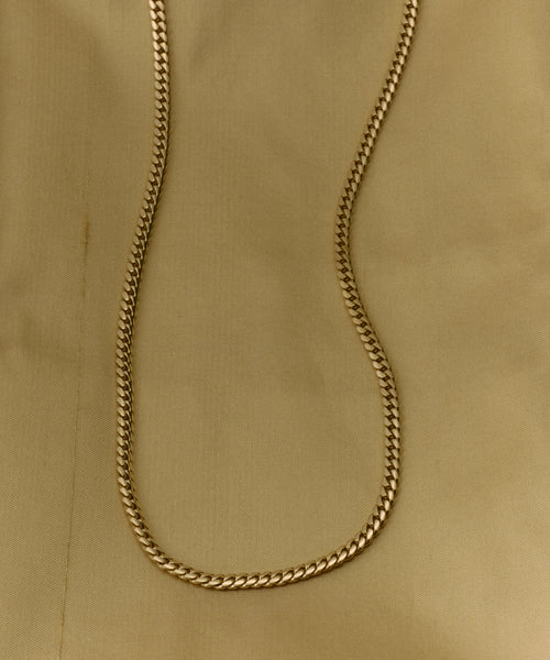 The Merit Chain, a 14k solid gold Cuban chain necklace, rests on beige fabric in a gentle curve. Its flat, interlocking links catch the light against a background featuring a faint seam and subtle vertical crease.