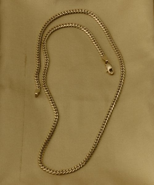 The Merit Chain, a 14k solid gold necklace with a lobster clasp, features medium thickness and a flat, interlocking curb link design. It's showcased on a light tan fabric background.