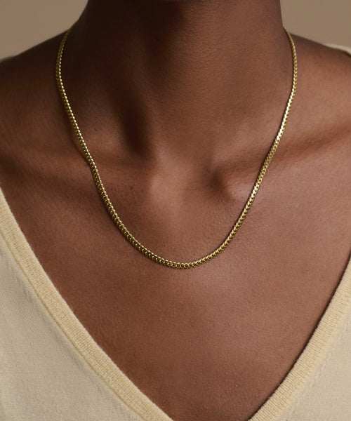 A person wearing a light beige V-neck top showcases the Merit Chain, a delicate 14k solid gold necklace, resting on their collarbone against a neutral background.