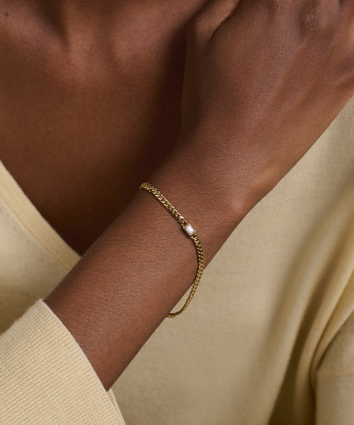 A close-up of a wrist wearing the Merit Emerald Cut Bracelet, featuring fine gold with a small silver accent. The wearer has on a light beige long-sleeved top against a neutral background.