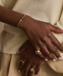 A close-up of crossed hands showcases the Merit Emerald Cut Bracelet and gold rings. The person wears a light yellow top, tan pants, and pale pink manicured nails, enhancing the elegant appeal of this fine jewelry look.