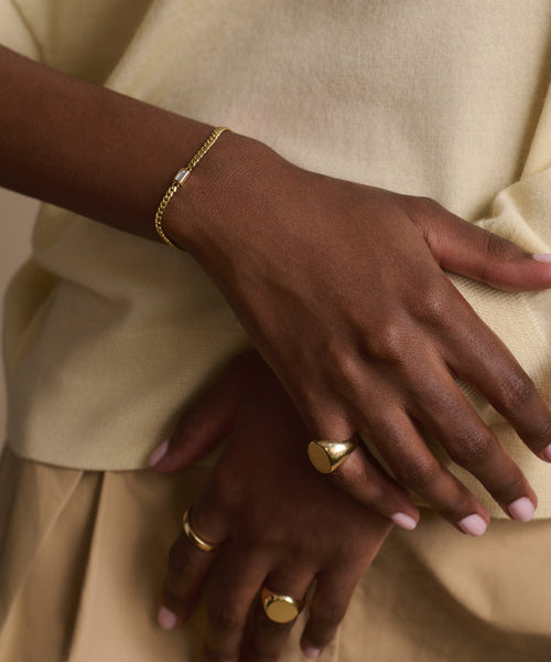 A close-up of crossed hands showcases the Merit Emerald Cut Bracelet and gold rings. The person wears a light yellow top, tan pants, and pale pink manicured nails, enhancing the elegant appeal of this fine jewelry look.