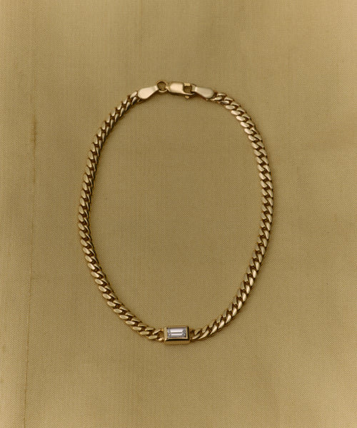 The Merit Emerald Cut Bracelet is a fine gold jewelry piece featuring a rectangular emerald-cut gemstone at its center, elegantly showcased on a light beige fabric background.