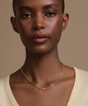 A person with short hair wears a light beige V-neck top and the Merit Emerald Cut Necklace, a simple 14k gold piece with a small emerald cut pendant, standing against a neutral beige background.