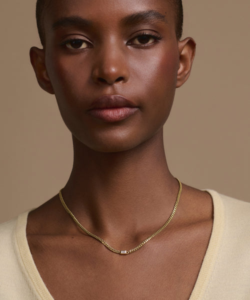 A person with short hair wears a light beige V-neck top and the Merit Emerald Cut Necklace, a simple 14k gold piece with a small emerald cut pendant, standing against a neutral beige background.