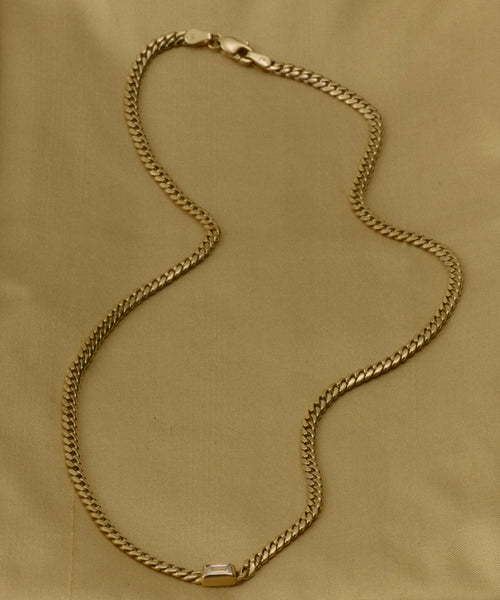 The Merit Emerald Cut Necklace features a 14k gold flat curb link design and a rectangular clasp, showcased against a gold-colored fabric background.