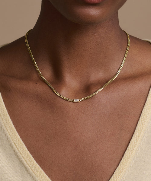 A person wearing the Merit Emerald Cut Necklace, featuring a 14k gold chain and small square pendant, shown from the shoulders up in a light beige V-neck top.